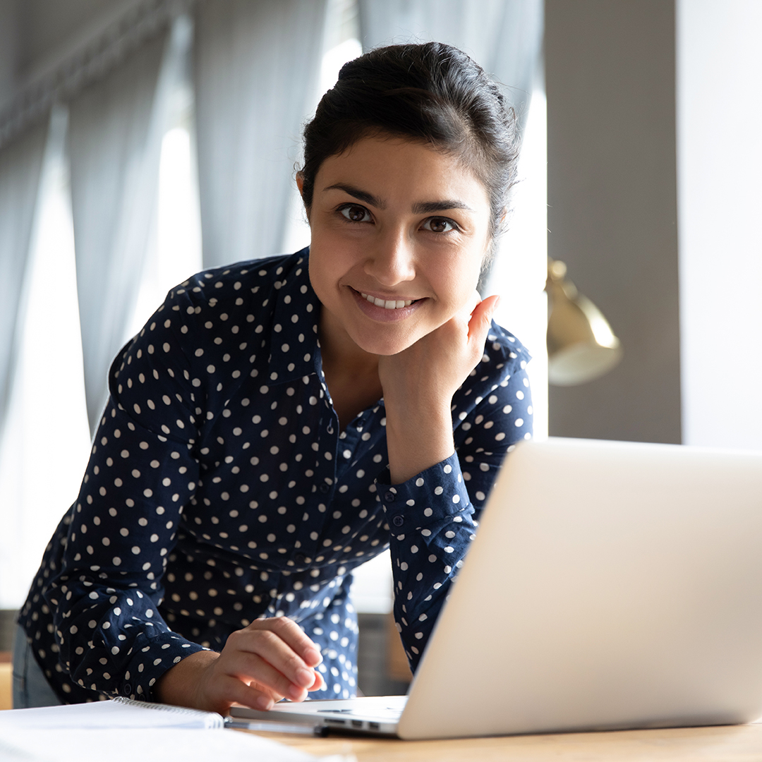 Portrait of smiling Indian girl stand at desk in living room working browsing Internet on modern laptop, profile picture of happy ethnic young woman busy surfing web, messaging typing on the gadget